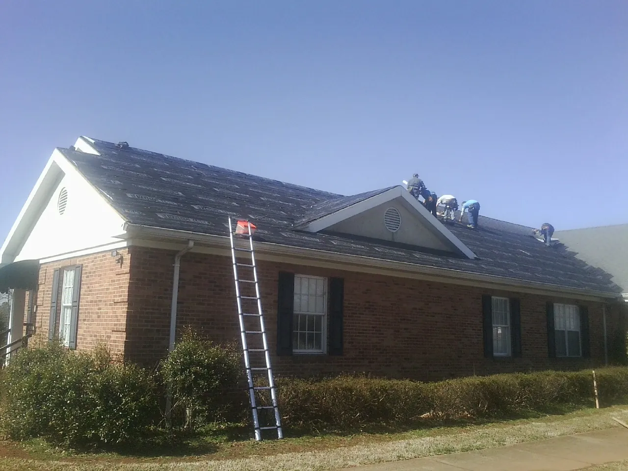 Crew tearing shingles off a brick ranch roof with a ladder set — Metal Roof Repair work by our crew in Round O