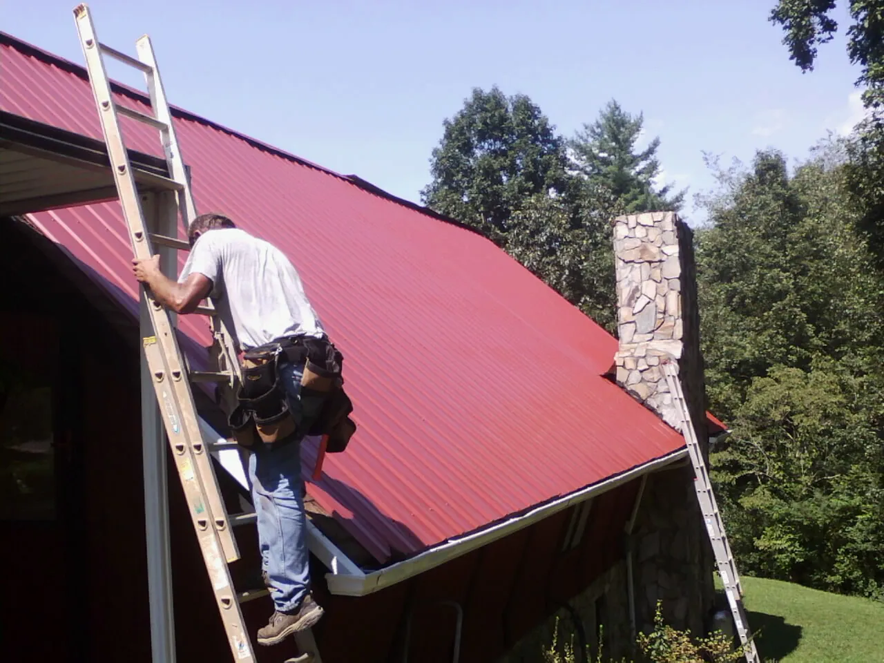 Roofer on a ladder beside a red metal roof and stone chimney — Soffit & Fascia Repair by Roofing Brothers in Round O